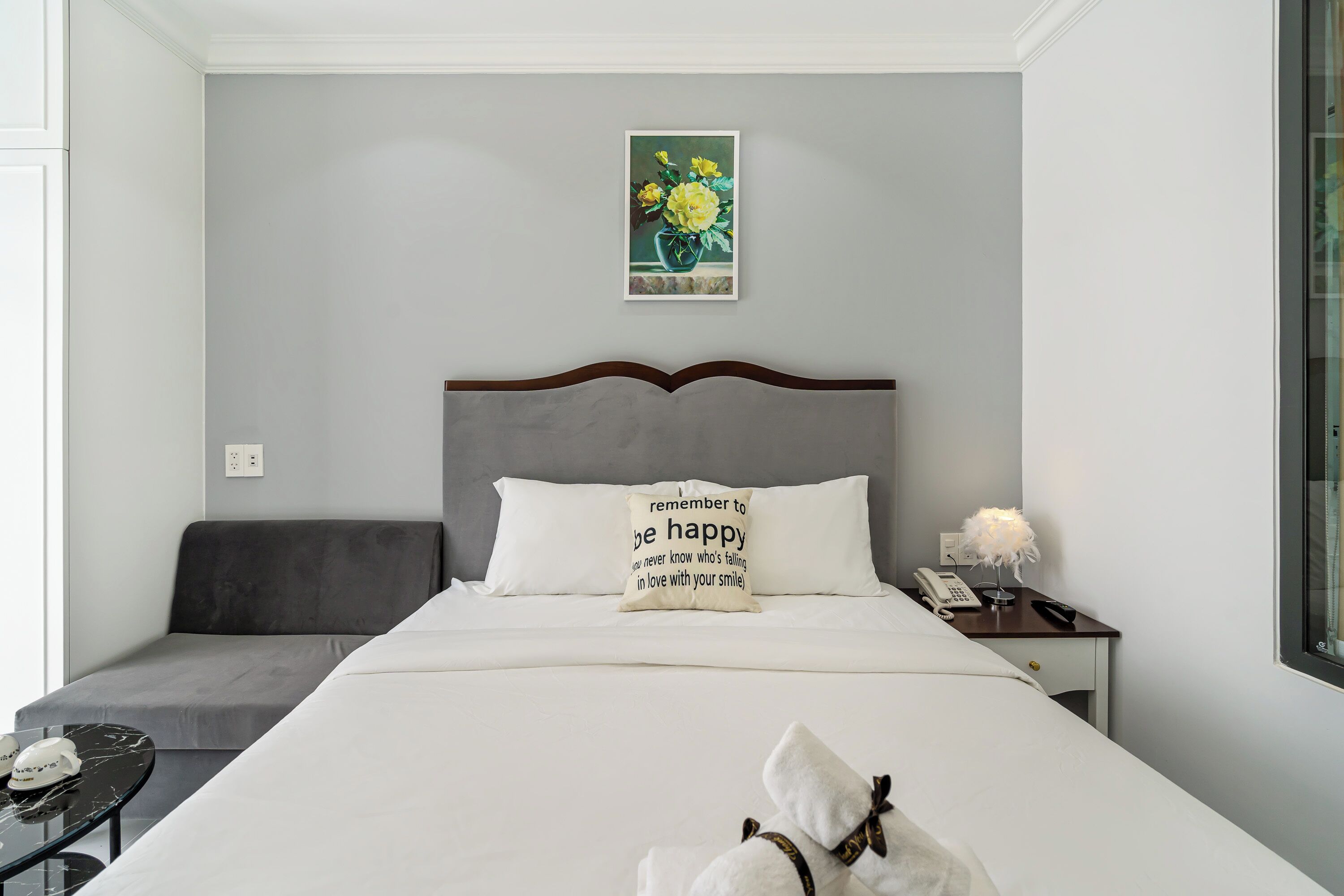 Ein ordentlich eingerichtetes Hotelzimmer mit einem Bett, einem Nachttisch mit Lampe, einem kleinen Tisch mit Telefon und einer Dekoration sowie einem Wandbild mit Blumen.
