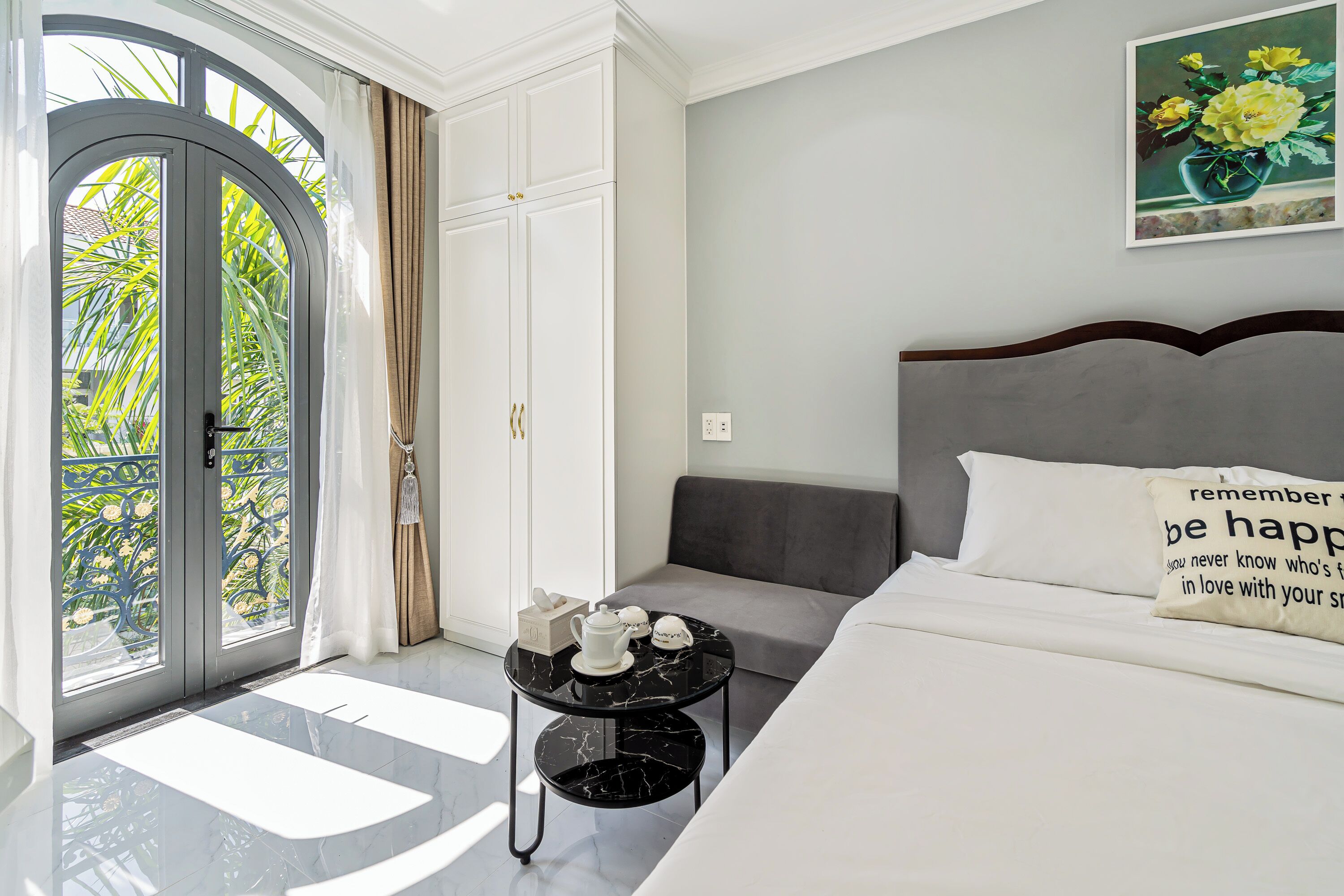 A hotel room with a bed, a small table with tea set, a grey sofa, and a painting on the wall.