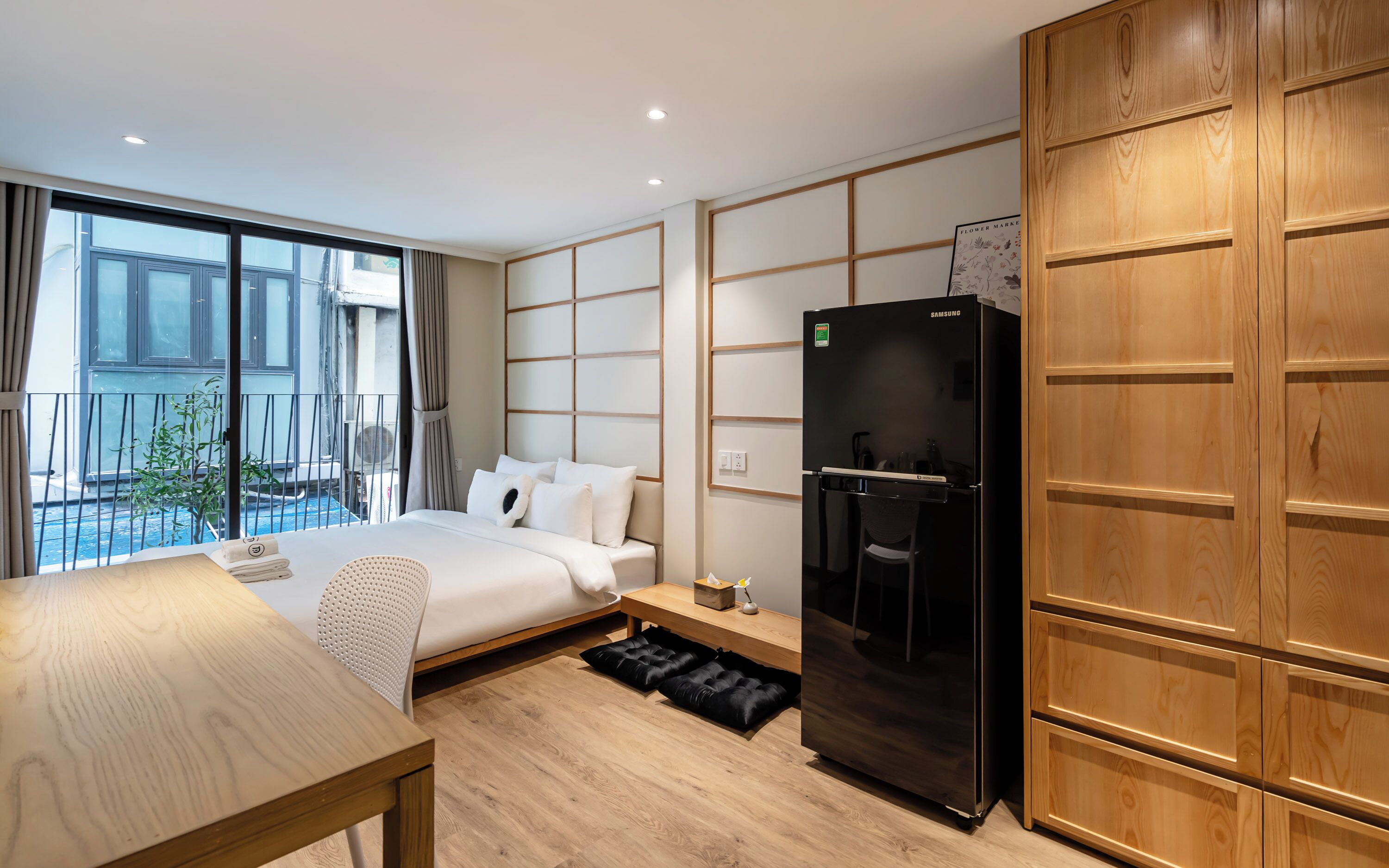 A modern hotel room with a bed, a wooden desk, a black refrigerator, and a view of the outside balcony.