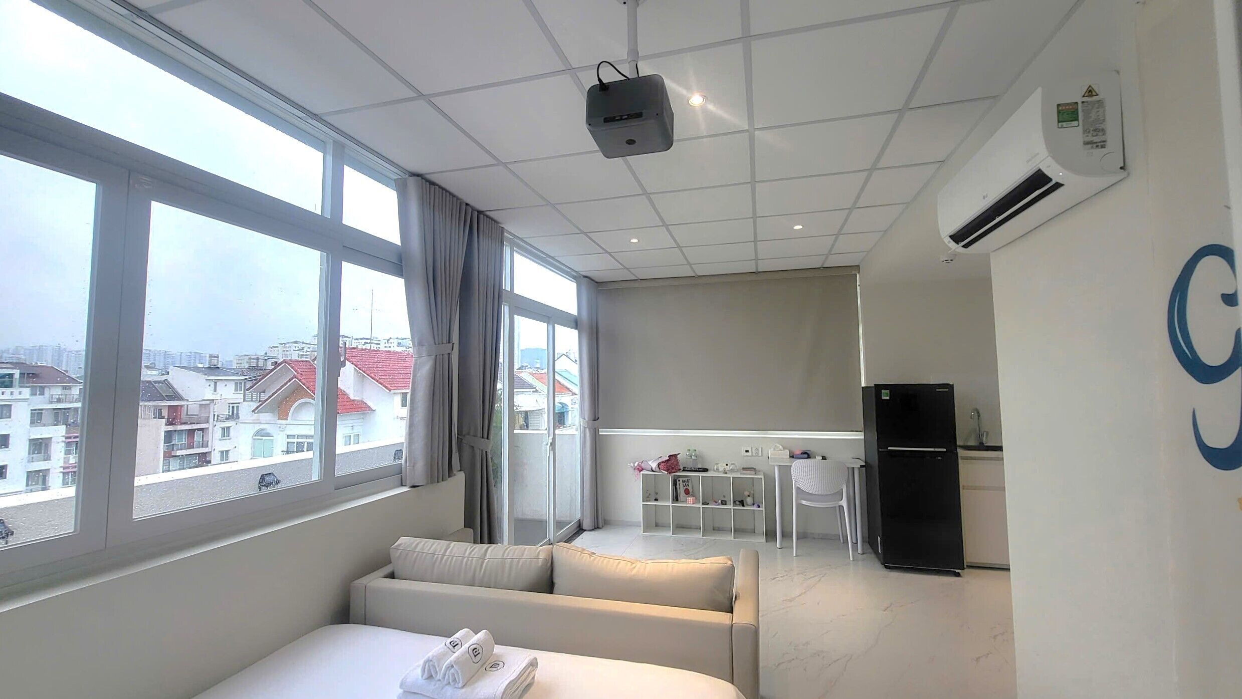 A hotel room with a bed, a sofa, a small table, a refrigerator, and a view of the city through large windows.