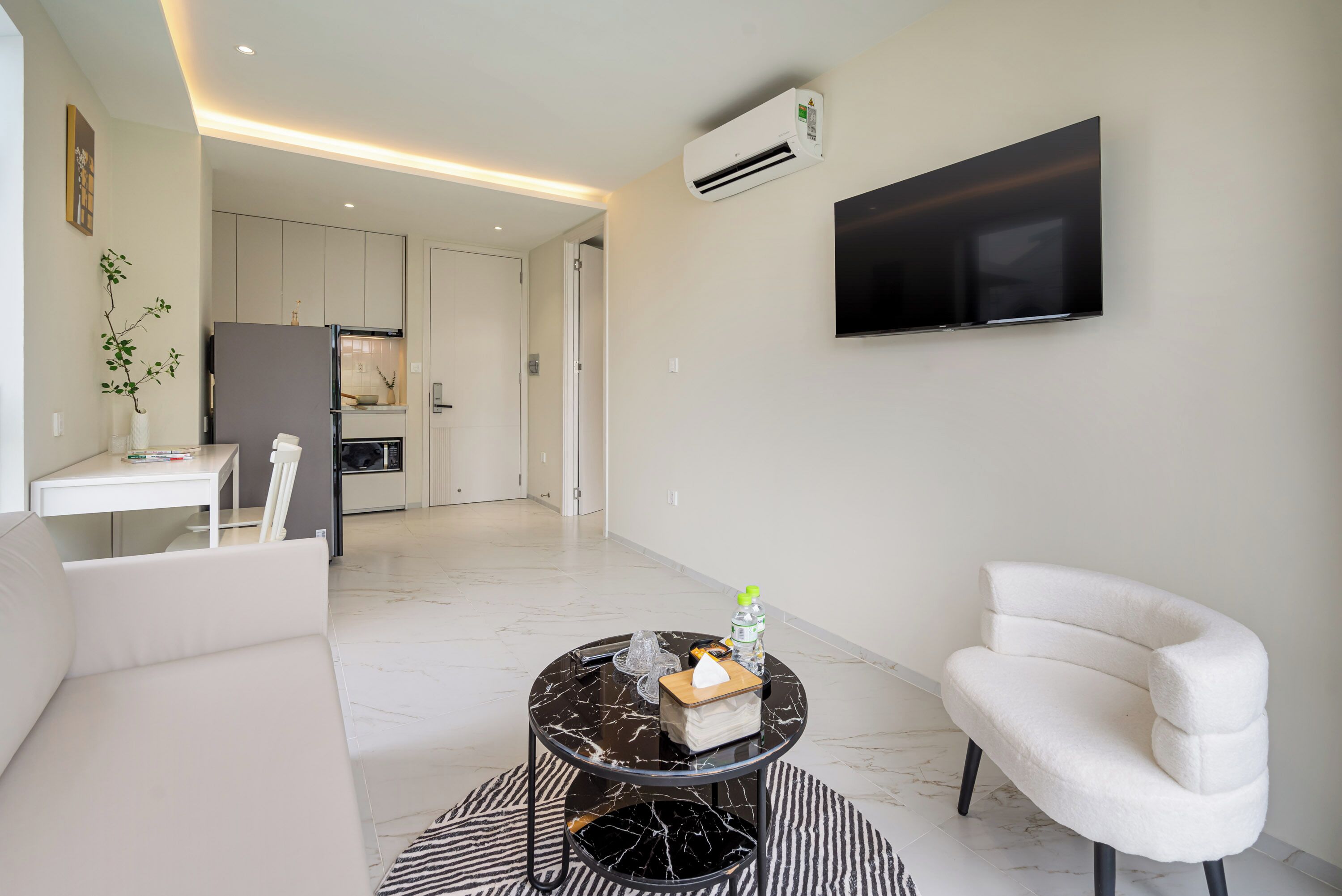Una sala de estar moderna con un sofá, un sillón blanco, una mesa de centro de mármol y un televisor de pantalla plana montado en la pared.