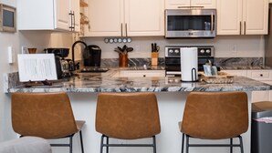 A modern kitchen with white cabinets, a stainless steel microwave, and a marble countertop. There are three brown bar stools and a grey sofa with a wooden frame.