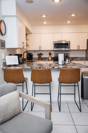 A modern kitchen with white cabinets, a stainless steel microwave, and a marble countertop. There are three brown bar stools and a grey sofa with a wooden frame.