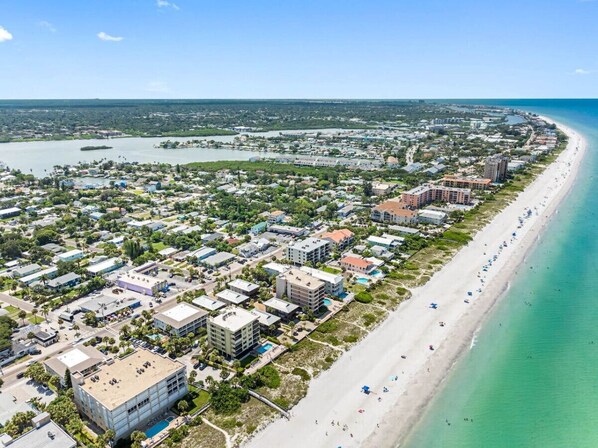 An aerial view of a coastal town with a long sandy beach, a marina, and a mix of residential and commercial buildings.