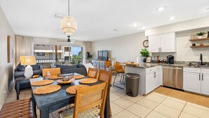 A modern kitchen with white cabinets, a dining area with a table and chairs, and a living room with a sofa and a TV.