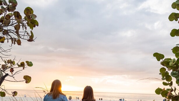 Two people walking on a sandy path towards a beach with a sunset in the background.