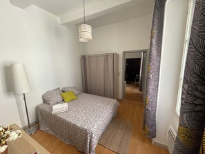 A bedroom with a bed, a floor lamp, a mirror, and a view of a hallway. - Appartement Centre-ville (Uzès)