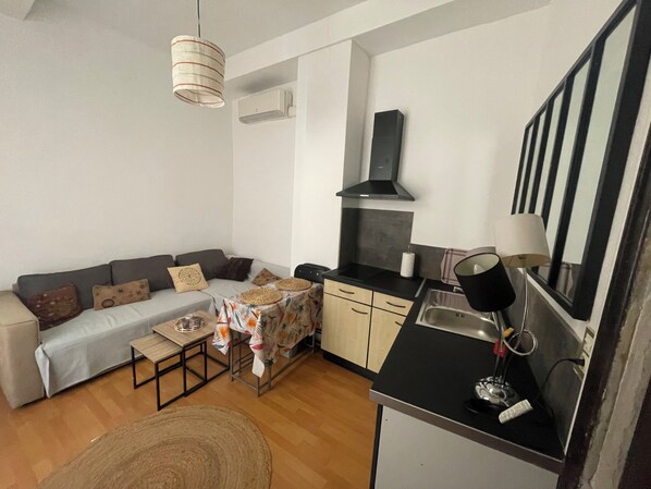 A compact living space with a sofa, a small table, a kitchen area with a sink and stove, and a ceiling fan. - Appartement Centre-ville (Uzès)