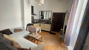 A compact living space with a sofa, a small table, a kitchen area, and a door leading to another room. - Appartement Centre-ville (Uzès)