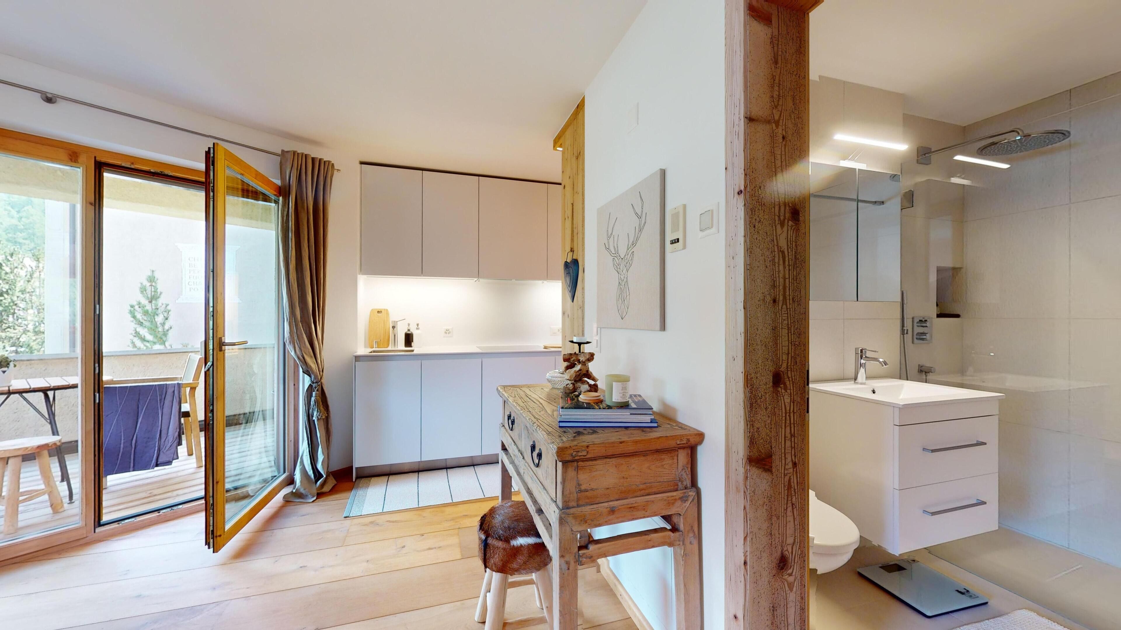 A modern bathroom with a shower, toilet, and sink. There is a wooden bench with a dog, a small table with books, and a window with curtains.