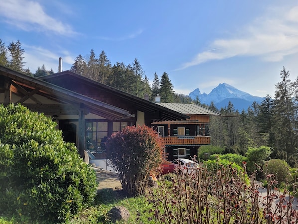 A wooden chalet with a balcony, surrounded by lush greenery and a mountain backdrop.
