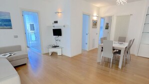 Een moderne woonkamer met een bank, een eettafel met stoelen, een tv op een dressoir, en uitzicht op de keuken door een open deur.