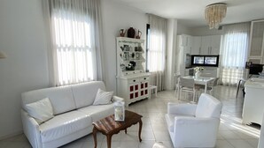 A modern living room with a white sofa, a wooden coffee table, and a white armchair. There is a glass cabinet with decorative items and a television in the background. - Villa Trieste Flexrent - Abyssinia (Riccione)