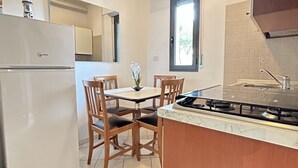 Une cuisine compacte avec un réfrigérateur blanc, une table à manger en bois avec quatre chaises, une petite plante en pot et une cuisinière à gaz.