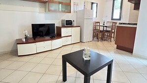 Une cuisine moderne équipée d’un réfrigérateur, d’un four à micro-ondes et d’une cuisinière. On y trouve un coin repas avec des chaises en bois et une petite table. Un téléviseur est posé sur un meuble, et des plantes en pot sont disposées sur des étagères.