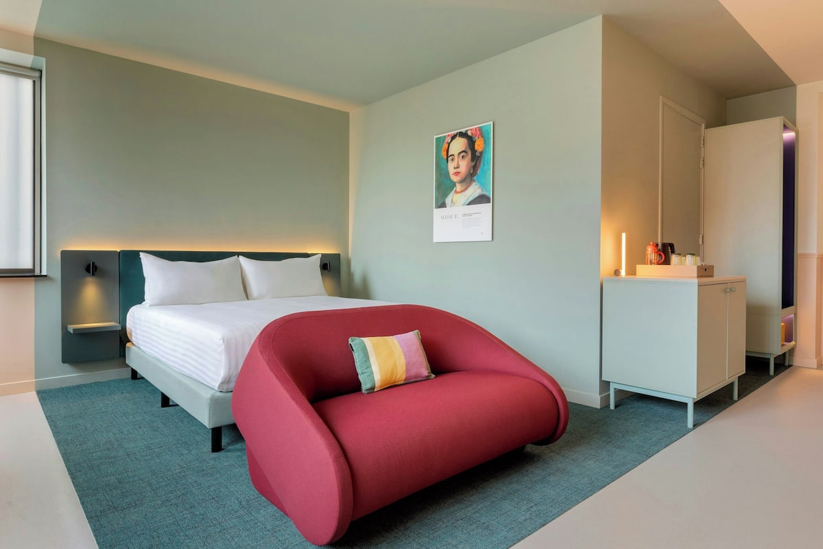 A modern bedroom with a bed, a red chair, a white dresser, and a wall-mounted shelf.