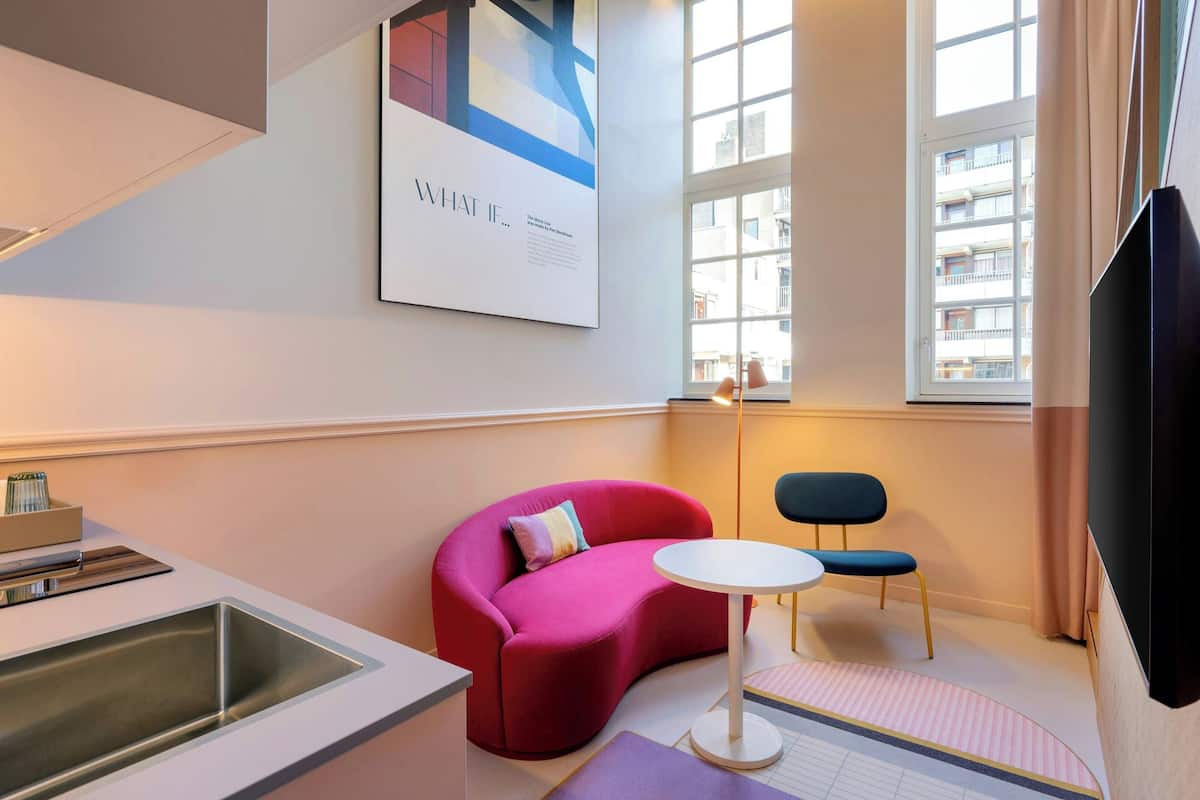A modern hotel room with a pink sofa, a small round table, a chair, a TV, and a large window with a view of buildings.