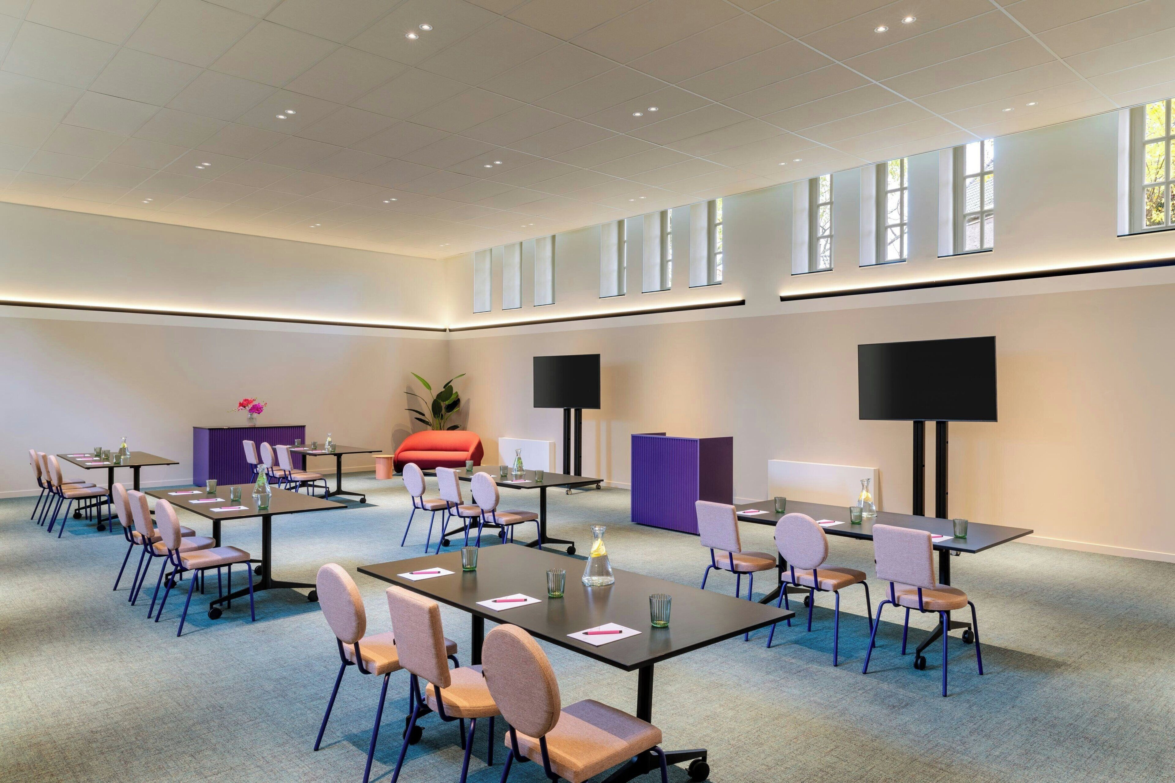 Une salle de conférence dotée de plusieurs tables et chaises, de grandes fenêtres et de deux grands écrans.