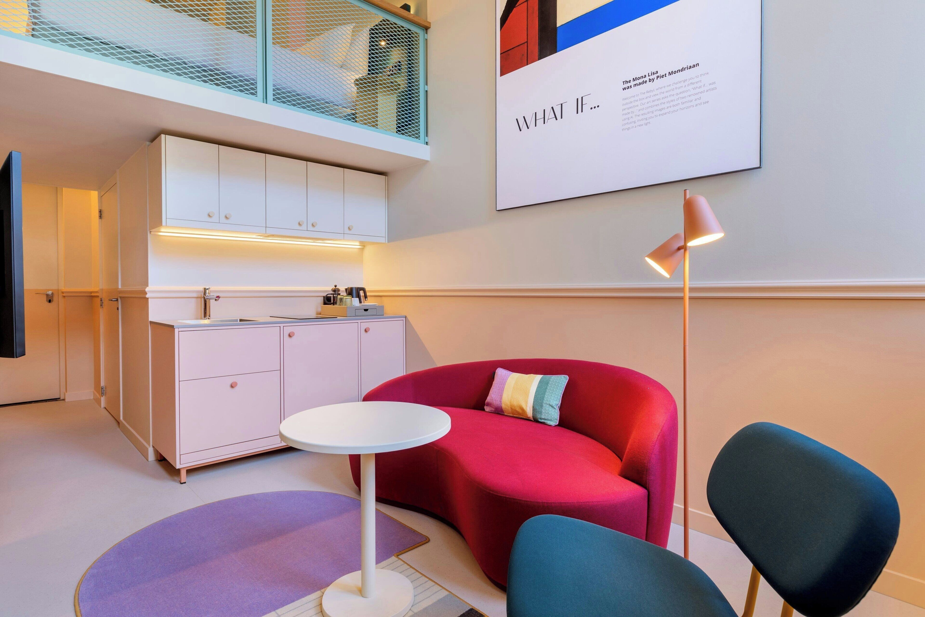 Ein modernes Interieur mit einem roten Sofa, einem weißen Tisch und einem blauen Stuhl. Es gibt eine Küchenzeile mit weißen Schränken und einer Arbeitsplatte. Ein gerahmtes Kunstwerk hängt an der Wand, und eine Stehlampe ist sichtbar.
