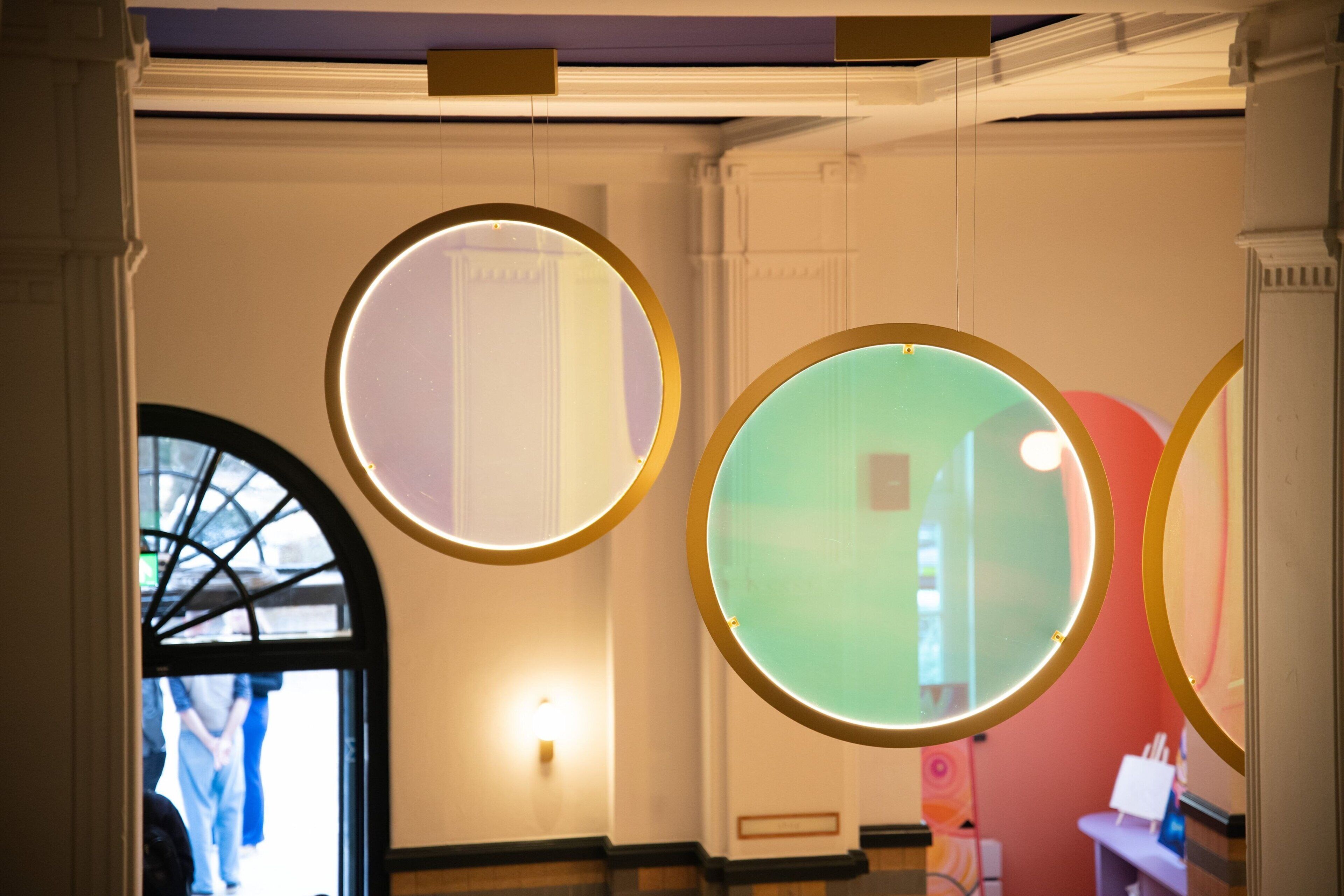 A hallway with three illuminated circular mirrors on the wall, a door with a glass panel, and a small table with a lamp.