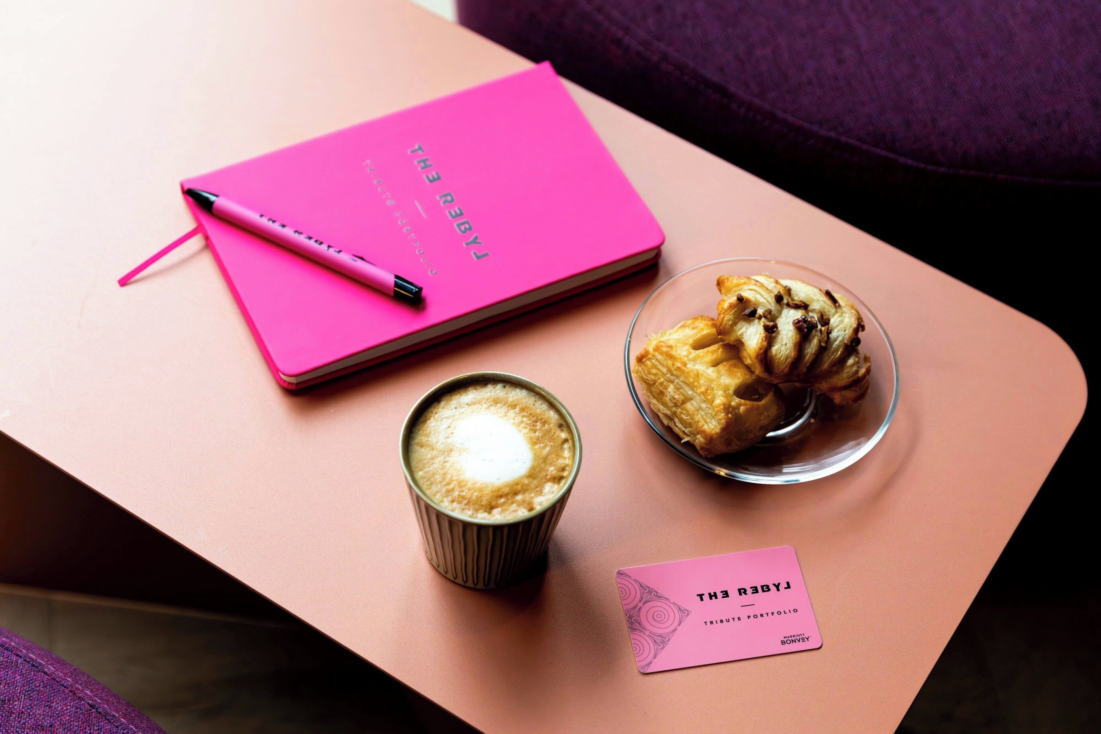 A pink notebook, a pen, a glass of coffee, a pastry, and a business card on a table.