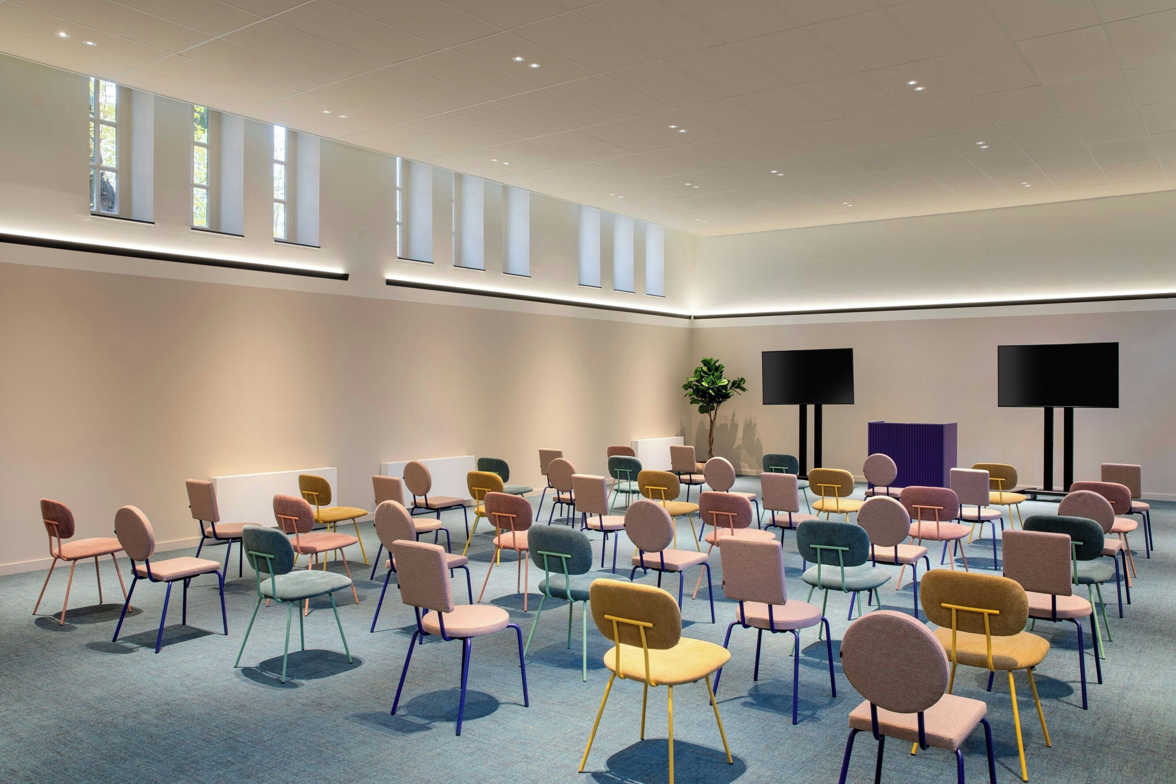 A conference room with rows of colorful chairs, a large window, and two black screens on the wall.