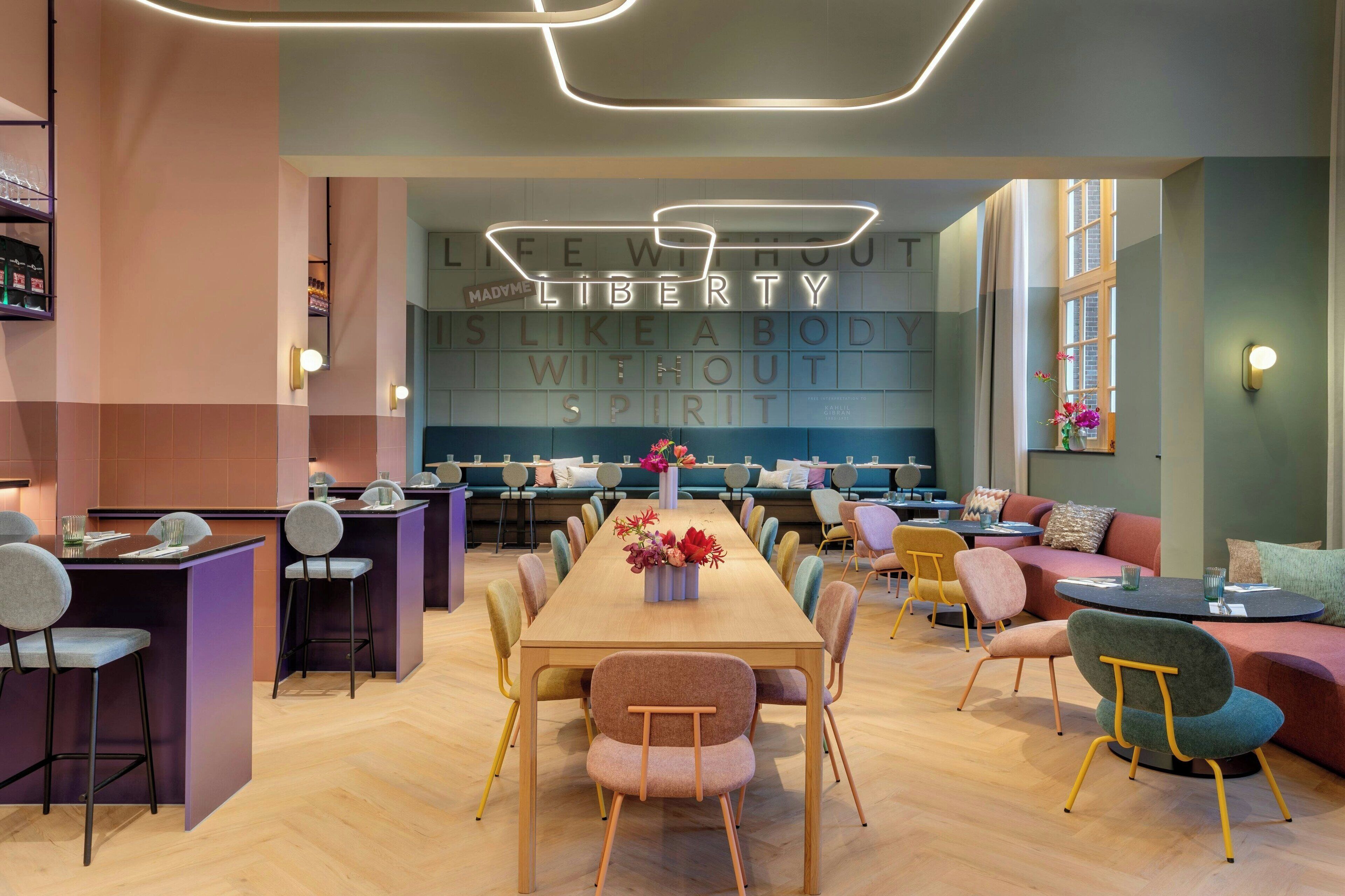 Een moderne lounge met een lange houten tafel, kleurrijke stoelen en een neonbord aan de muur.