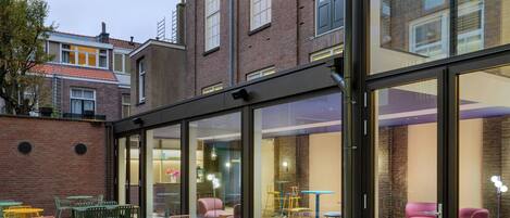 A modern outdoor seating area with tables and chairs, a glass door entrance, and a brick building in the background.