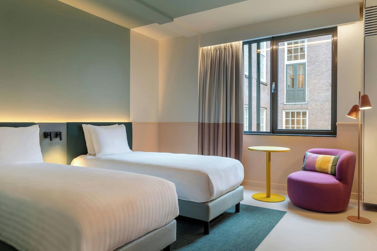 A hotel room with two beds, a small round table, a purple chair, and a window with curtains.