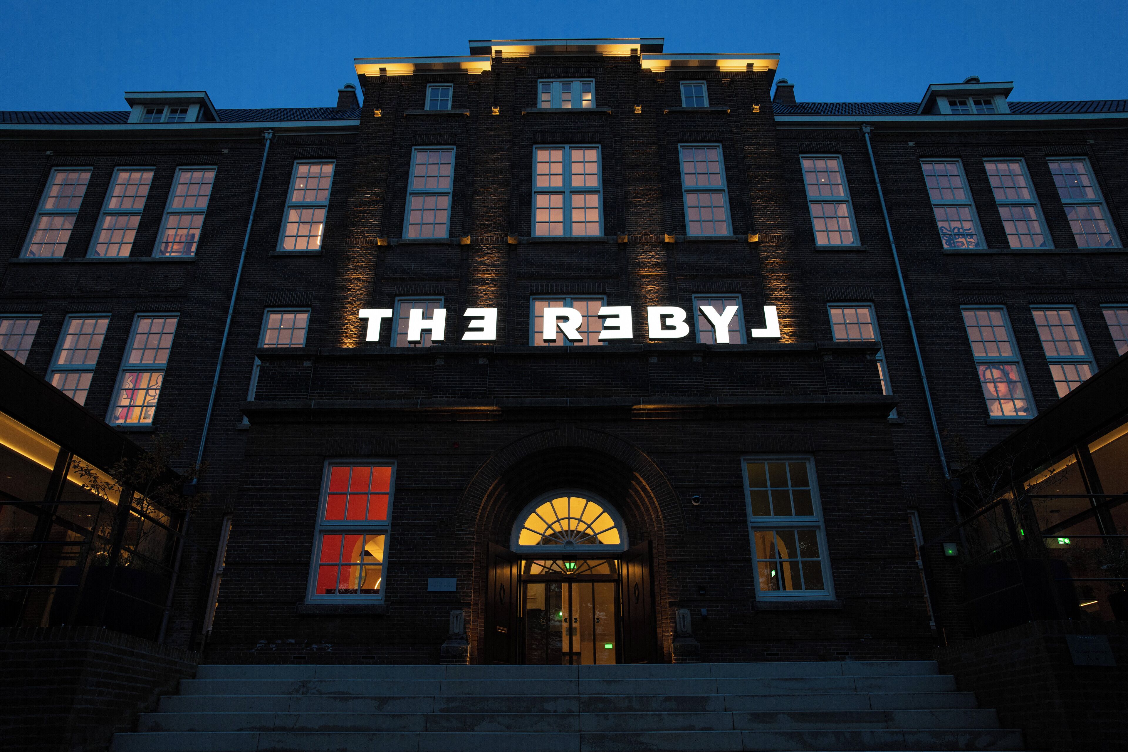 A building with "THE REBY" illuminated signage, a brick facade, and a central entrance with a glass door.