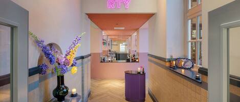 A hotel corridor with a neon sign, a vase with flowers, a reception desk, and a herringbone floor pattern.
