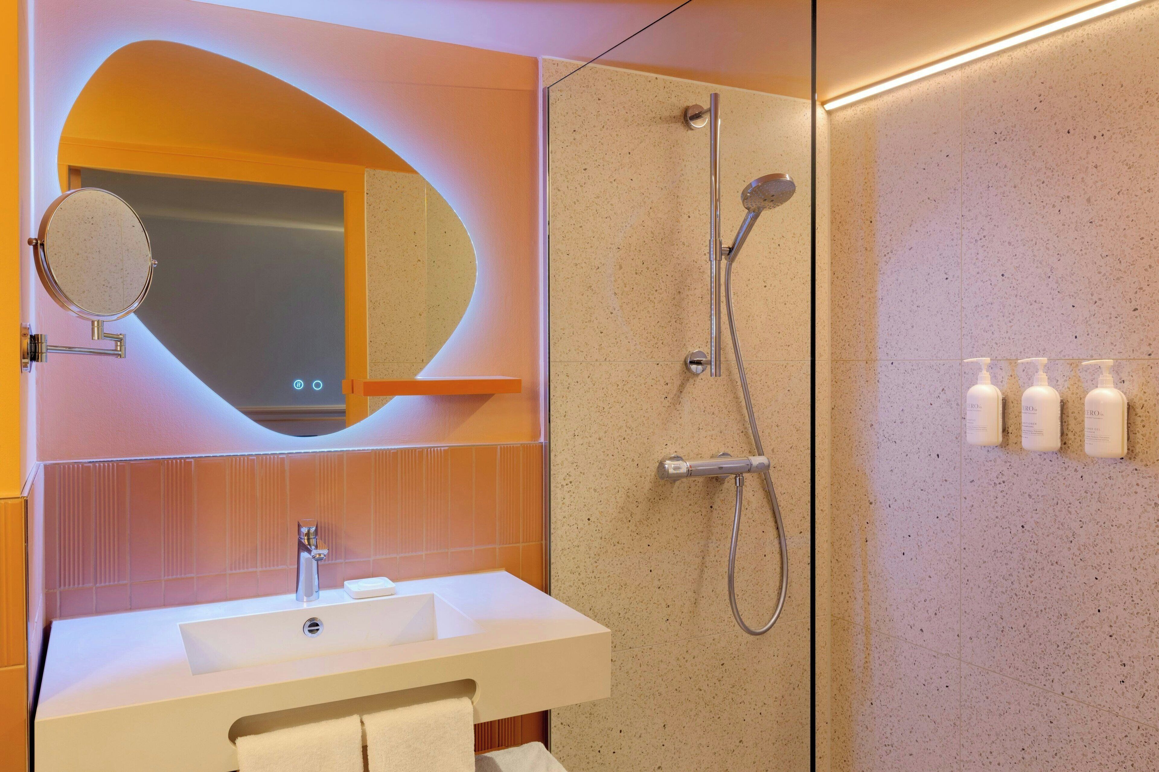 Ein modernes Badezimmer mit einem weißen Waschbecken, einem runden Spiegel, einer Dusche inklusive Handbrause und drei Toilettenartikeln an der Wand.