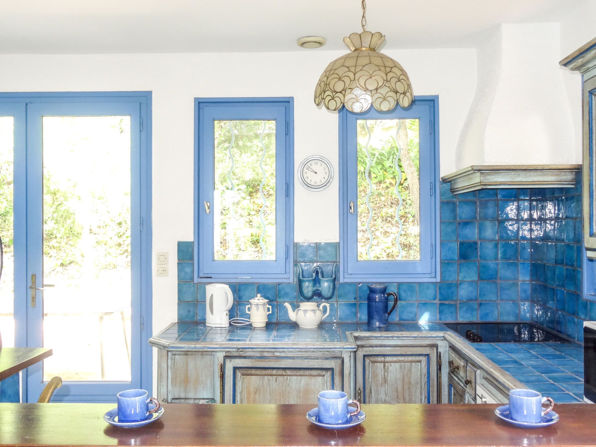 Eine Küche mit blau gefliesten Wänden, zwei Fenstern mit blauen Rahmen, einer Wanduhr und einem Holztisch mit blauen Tassen.