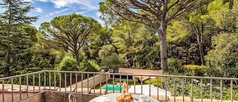 A round table with white chairs and a metal railing, surrounded by lush greenery and trees.