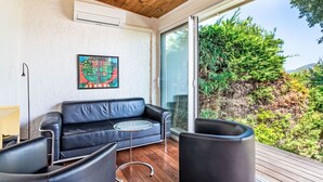 Una sala de estar moderna con un sofá de cuero negro, una mesa de centro de vidrio y dos sillones negros. Un cuadro colorido adorna la pared, y hay un amplio ventanal con vistas a la vegetación.