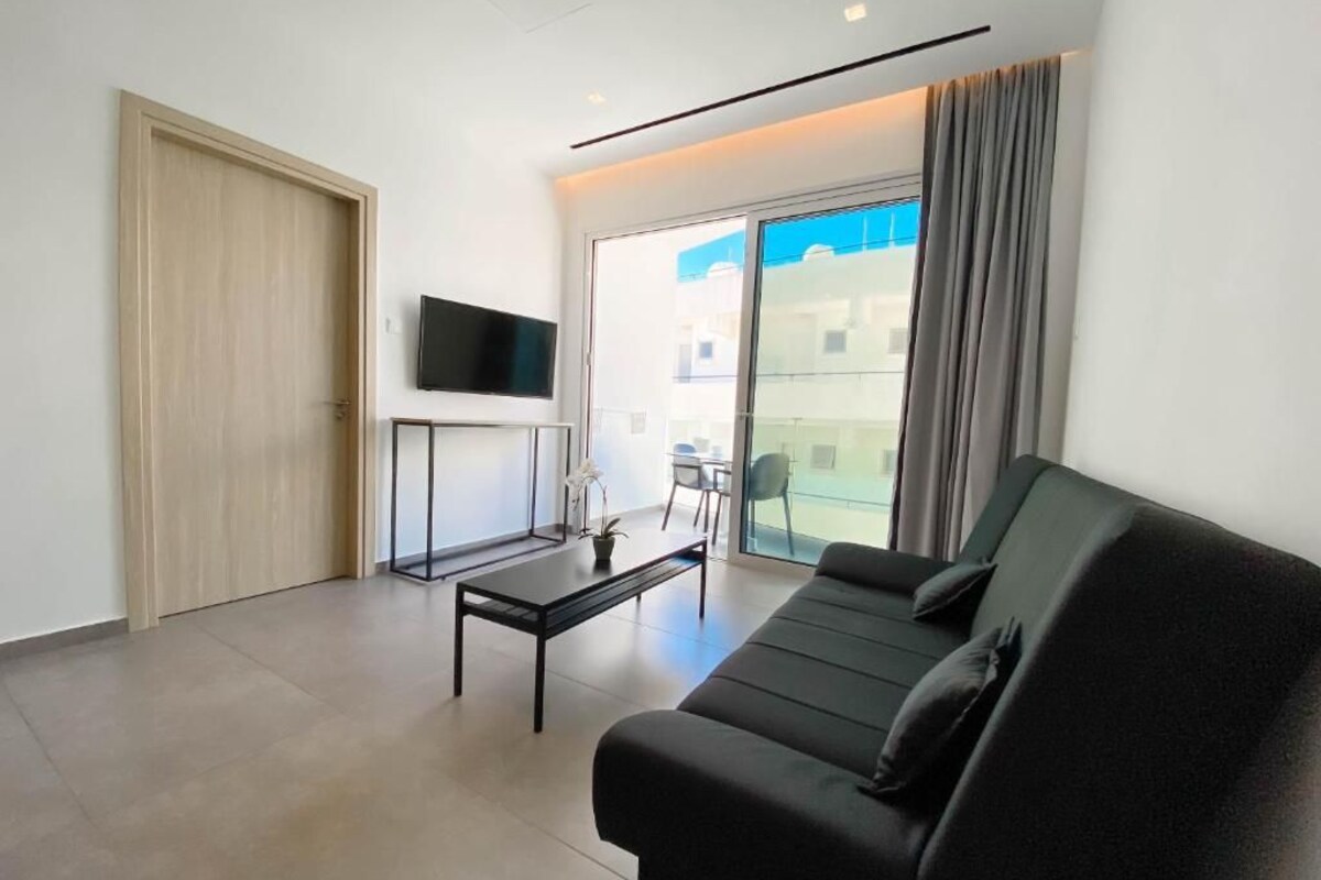 A modern living room with a black sofa, a coffee table, a flat-screen TV, and a sliding glass door leading to an outdoor area.