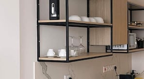 A kitchen with a black and wooden shelving unit holding white dishes, glasses, and a microwave. - Caramela Luxury Apartments (Ayia Napa)