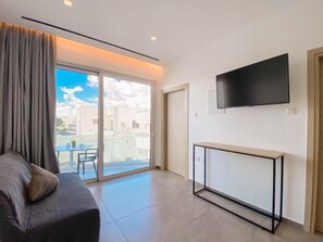 Ein modernes Wohnzimmer mit einer Couch, einem an der Wand befestigten Fernseher, einer Schiebeglasdoor zum Balkon und einem kleinen Tisch.