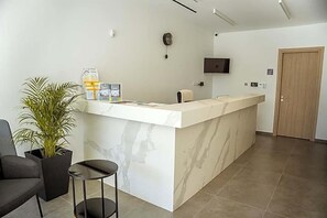 A reception area with a marble counter, a potted plant, a black chair, and a television mounted on the wall. - Caramela Luxury Apartments (Ayia Napa)