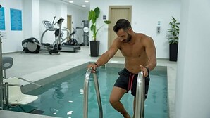 A man in black shorts with red trim stepping out of a swimming pool in a modern indoor facility with exercise equipment in the background. - Caramela Luxury Apartments (Ayia Napa)