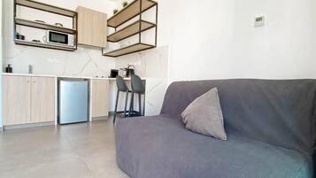 Una cocina moderna con microondas integrado, cafetera y refrigerador pequeño. En la sala de estar, hay un sofá gris con un cojín y una silla.