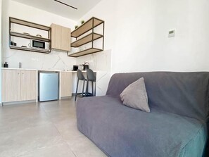 A modern kitchen with a built-in microwave, a coffee machine, and a small refrigerator. There is a grey sofa with a cushion and a chair in the living area. - Caramela Luxury Apartments (Ayia Napa)