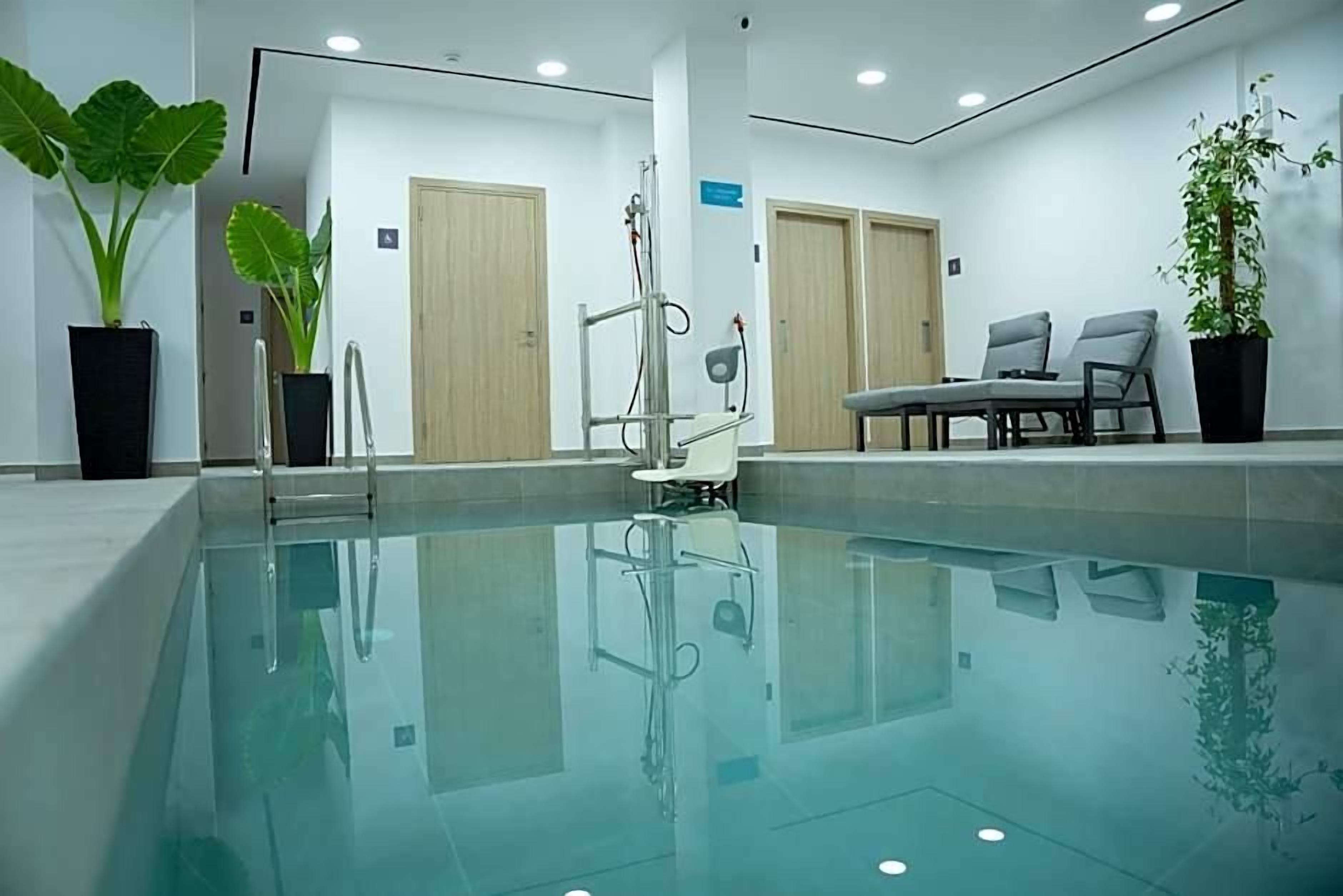 Una piscina interior moderna con paredes de vidrio transparente, puertas de madera y plantas en macetas.