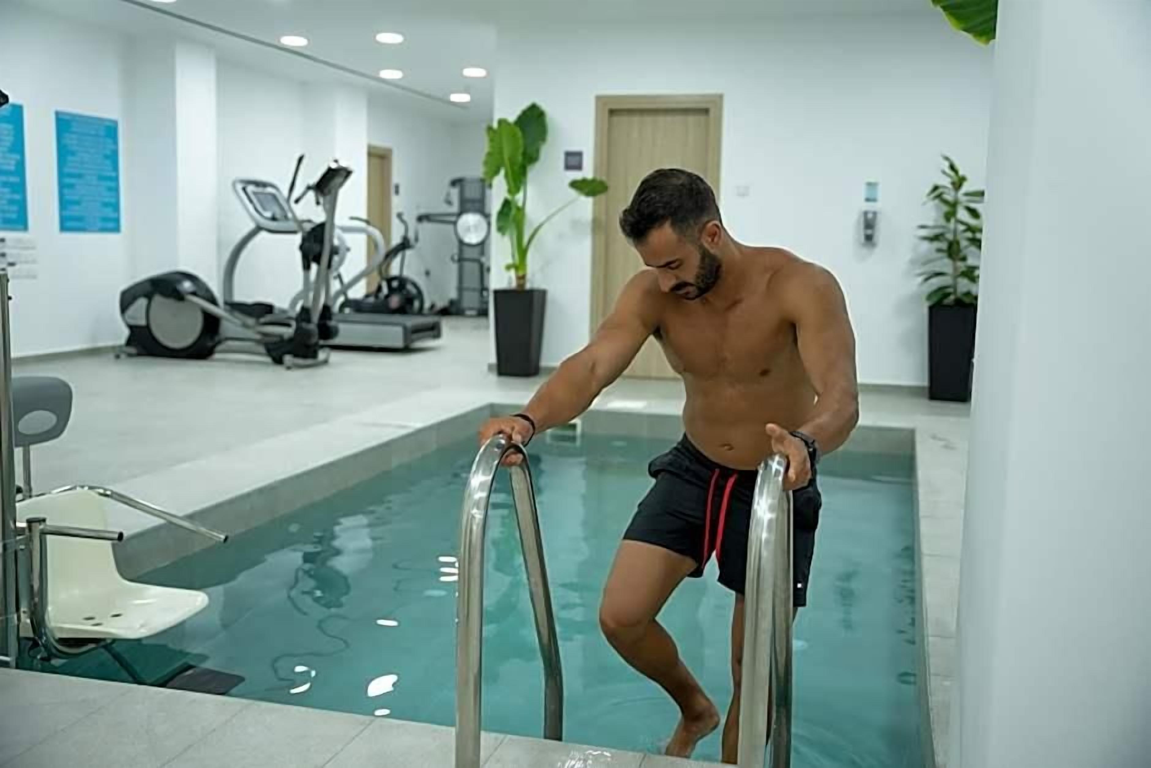 Un homme, vêtu d’un short noir orné d’une bande rouge, sort d’une piscine dans un complexe intérieur moderne, avec des équipements de musculation en arrière-plan.