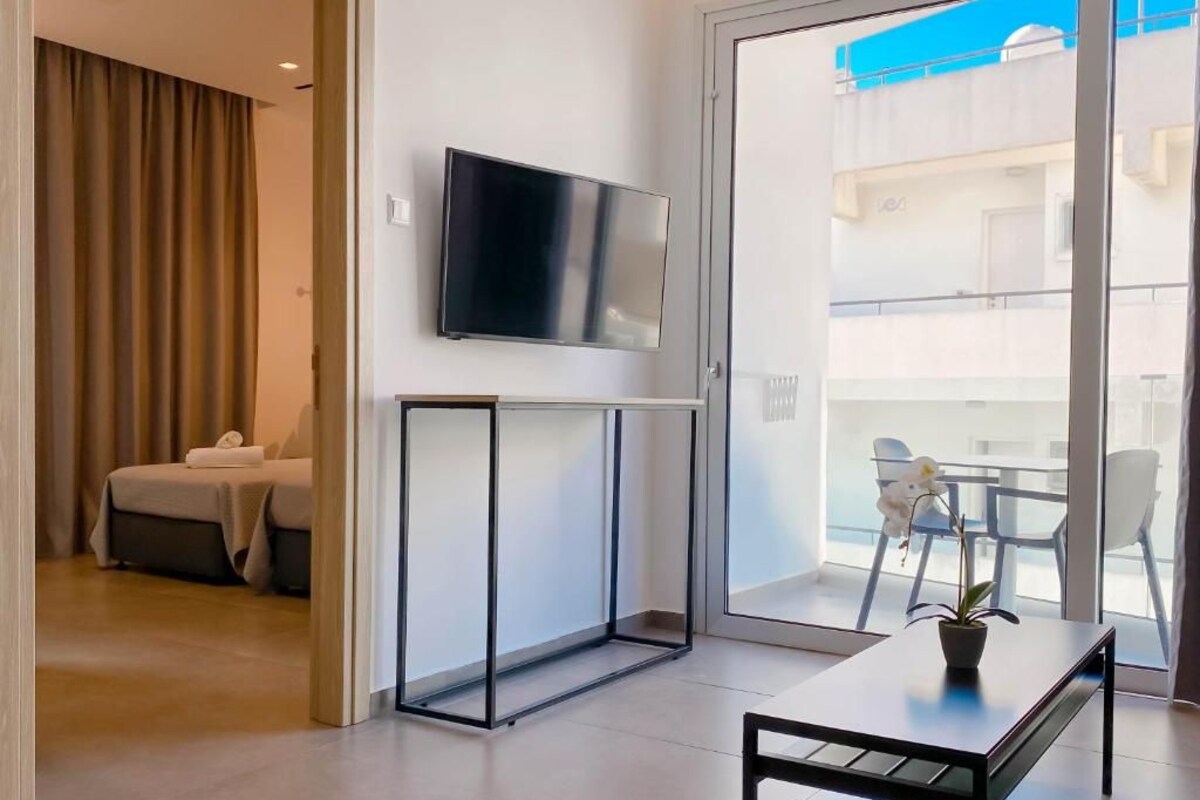 Een moderne hotelkamer met een flatscreen televisie aan de muur, een balkon met uitzicht op het gebouw en een zithoek met een tafel en stoelen.