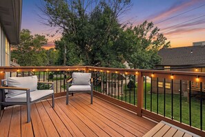 Une terrasse en bois avec deux chaises blanches dotées de coussins, une balustrade ornée de guirlandes lumineuses et une vue sur un jardin avec des arbres et une maison.