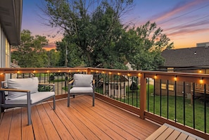 Una terraza de madera con dos sillas blancas acolchadas, una barandilla con luces y vistas a un jardín con árboles y una casa.