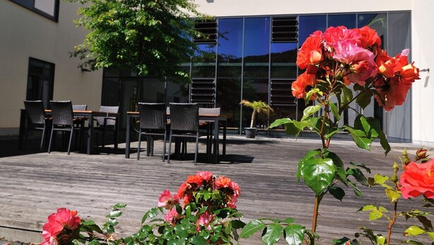 Un patio al aire libre con cubierta de madera, un área de estar con sillas y mesas, y un edificio con grandes ventanales al fondo.