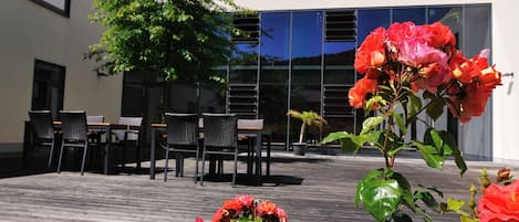 An outdoor patio with a wooden deck, a seating area with chairs and tables, and a building with large windows in the background.