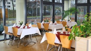 Un restaurante con mesas preparadas para comer, ventanas amplias y plantas de interior.
